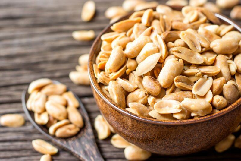 A wooden table with a large brown bowl of roasted peanuts, and a spoonful of them