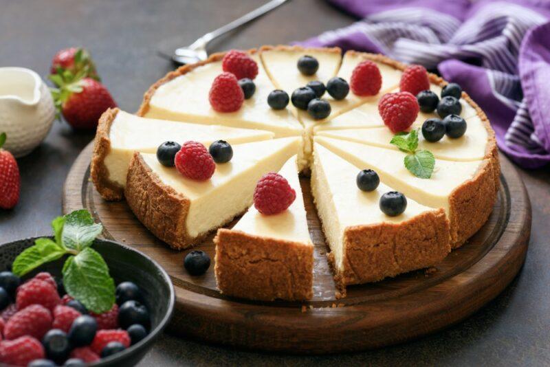 A wooden board with a large cheesecake that's been cut into many pieces with fruit on the top