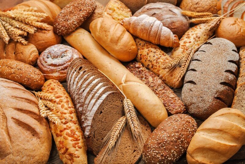 A wooden table with many different loaves of bread. One of the loaves has been sliced and there are also ears of wheat among the loaves.