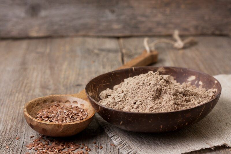 A large bowl of ground flaxseed, next to a wooden spoon that contains whole flax seeds