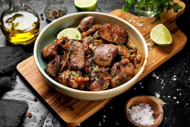 A wooden board with a bowl of cooked chicken liver. There are ingredients surrounding it, including salt, olive oil, and limes