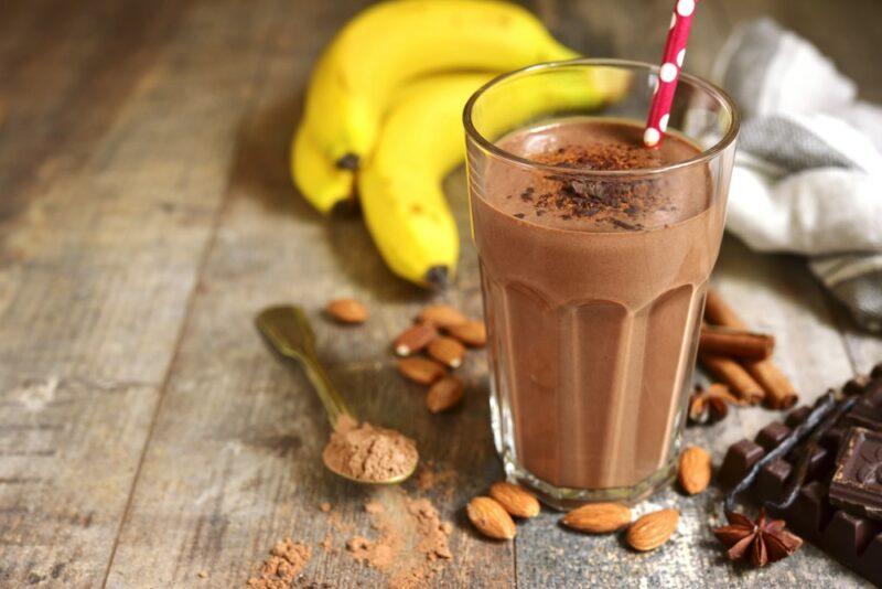 A tall glass containing a chocolate banana protein smoothie on a wooden table, in front of protein powder, nuts, and bananas