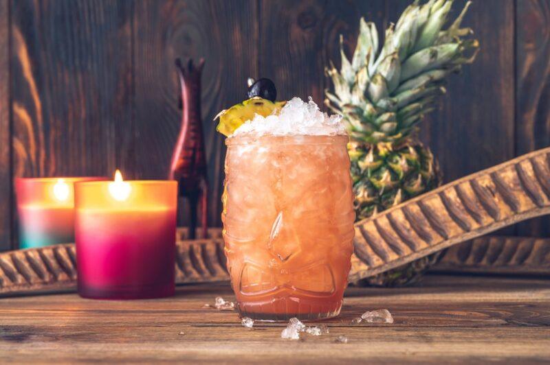 A large glass containing a jungle juice cocktail with plenty of ice, in front of a pineapple and some candles