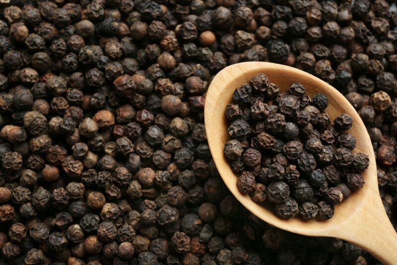 A large pile of black peppercorns, with a wooden spoon that's filled with even more peppercorns