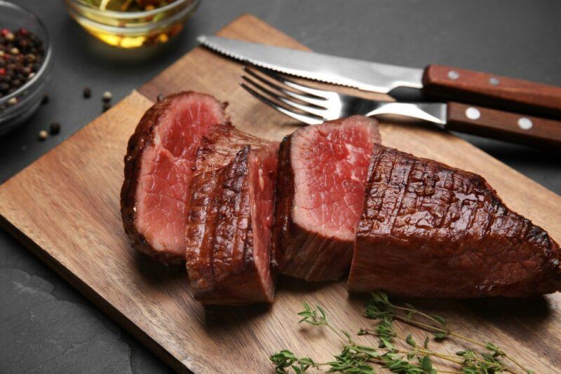 A wooden board with a large tenderloin that's been cut into pieces, next to a carving knife and fork, with herbs and seasonings nearby.