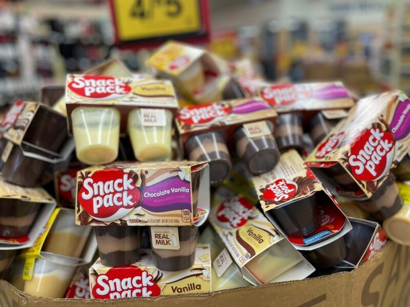 A large pile of Snack Pack desserts in a grocery store, with the shelves and price signs blurred out in the background