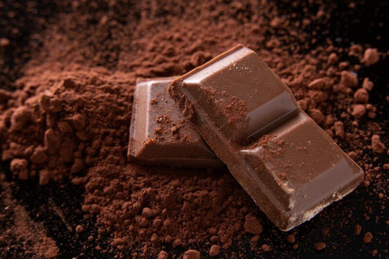 A pile of cocoa powder on a black table, with a few squares of chocolate on top.