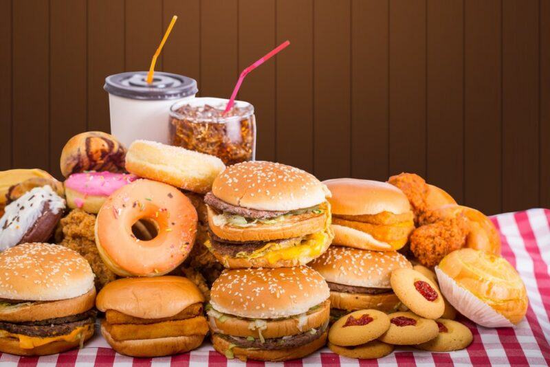 A red and white checkered cloth, piled high with different types of fast food, including many burgers, donuts, fried chicken, and treats, plus two cups of soda