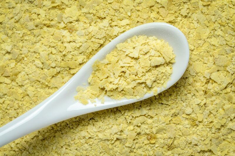 Light colored nutritional yeast in a pile with a white spoon that contains more of the yeast