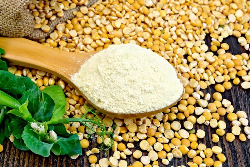 A wooden table with a large pile of split peas and a wooden spoon containing pea protein powder