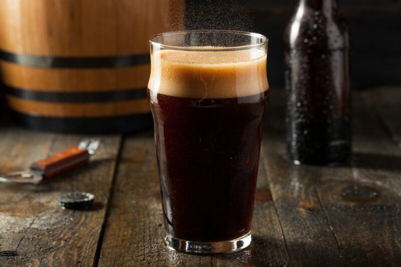 A wooden table with a large pint of hard root beer, next to a bottle opener, a barrel, and a bottle