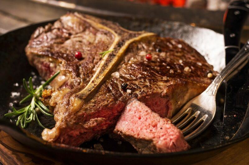 A black plate with a large porterhouse steak, a fork, and a little rosemary