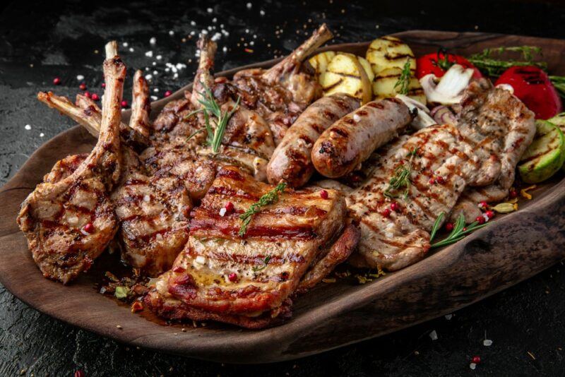 A large wooden tray with many types of cooked meat, plus cooked potatoes, tomatoes, and zucchini.