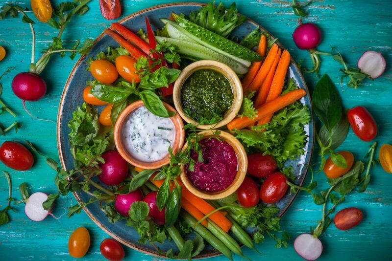 A large plate on a teal table, with a large variety of sliced and whole veggies, along with three small bowls of dip