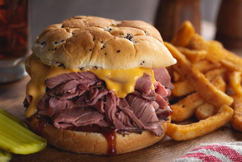 A large roll that is jam packed with sliced beef with a little mustard or melted cheese near the top. There are some fries in the background.