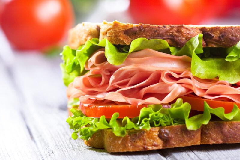 An impressive sandwich on a white table filled with lettuce, tomatoes, and ham, with some tomatoes in the background