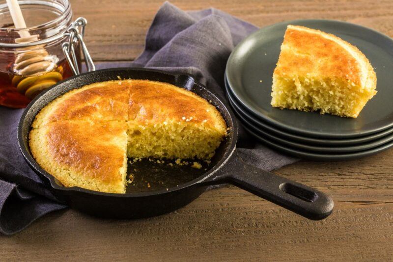 A skillet filled with cornbread, with one piece cut out, next to a stack of black plates, one of which contains a piece of cornbread