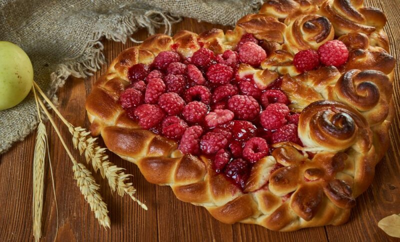 A large sweet pirog in the shape of a heart, with many raspberries, next to a couple of pieces of wheat