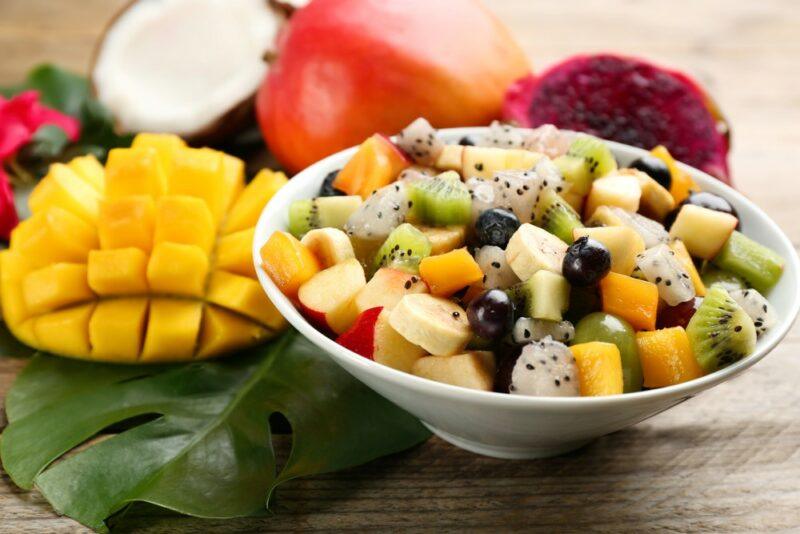 A large white bowl containing many pieces of fresh fruit, including dragonfruit and apples, next to some mangoes, coconut, and dragonfruit