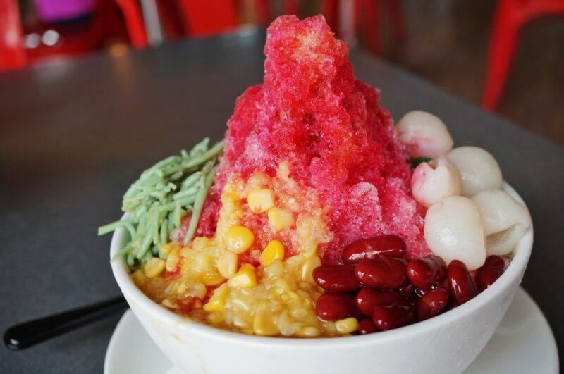 A white bowl containing a dessert called ais kacang, which is surrounded by plenty of toppings, including red beans