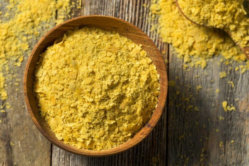 A wooden bowl filled with nutritional yeast, with a spoon of the yeast next to it and some sprinkled on the wooden table