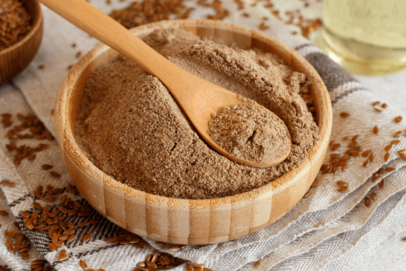 A large brown bowl containing ground flaxseeds with a wooden spoon resting on top