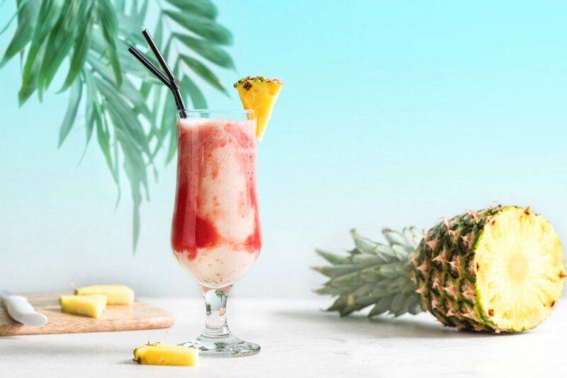 A tall lava flow cocktail in front of a light blue background with a pineapple next to it