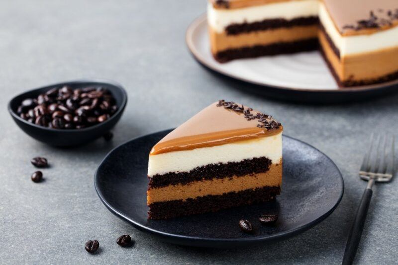 A black plate with a piece of layered caramel and coffee cake, with the rest of the cake and a bowl of coffee beans in the background