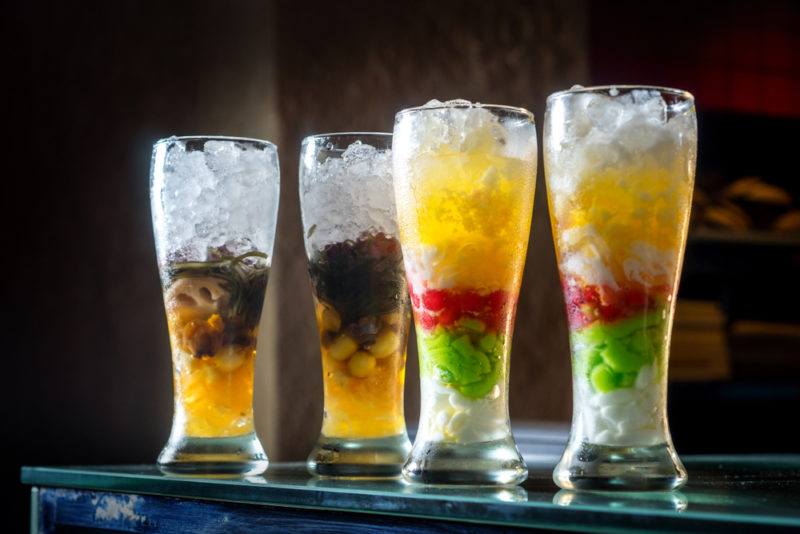 Four glasses of a layered Vietnamese dessert called che, with ice or gelatin cubes on the top