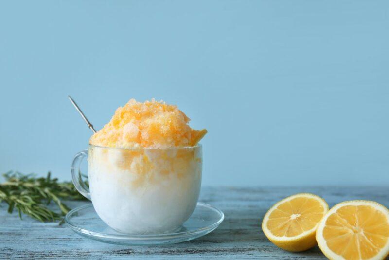 A glass mug containing a lemon lime snow cone, next to two lemon halves and some herbs