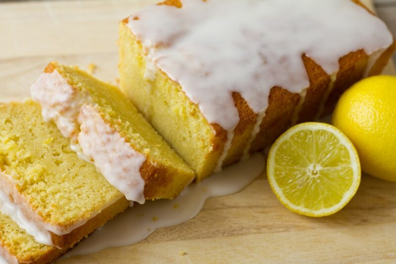 A light wooden table with a lemon pound loaf with icing, next to a fully lemon and a lemon half