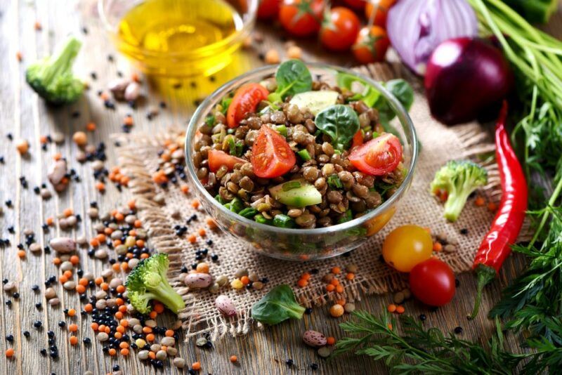 A dish filled with a lentil salad, with more lentils on the table, along with many different fresh ingredients