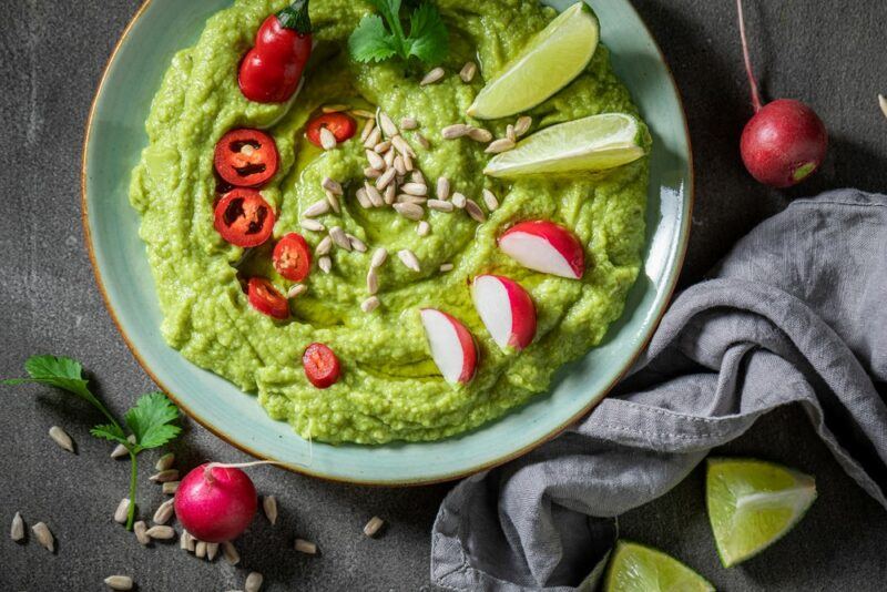 A light blue dish containing an avocado based dip with radishes, tomatoes, limes and sunflower seeds as garnishes