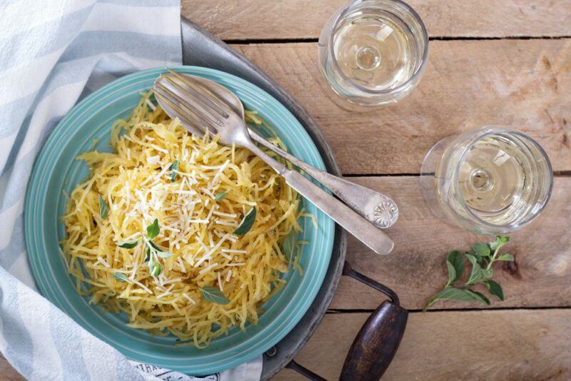 A light blue dish of spaghetti squash with cheese, next to a knife and fork and glasses