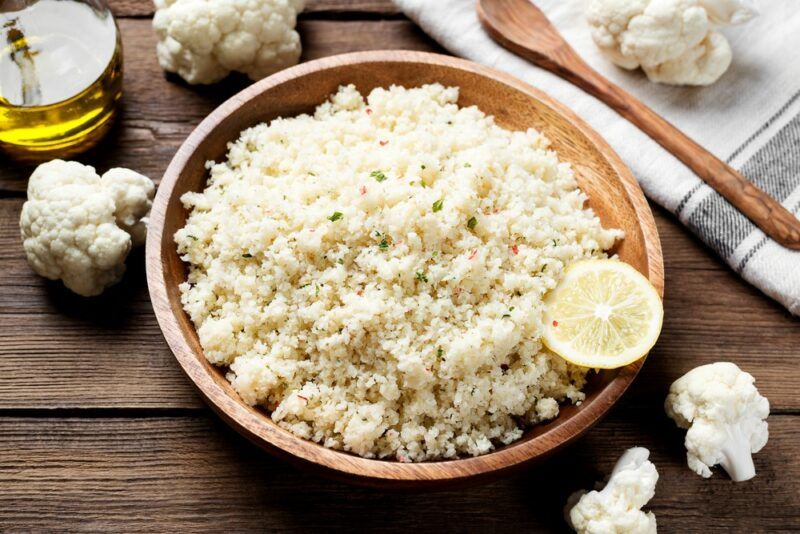 A light brown bowl filled with cauliflower rice, with cauliflower florets on the table