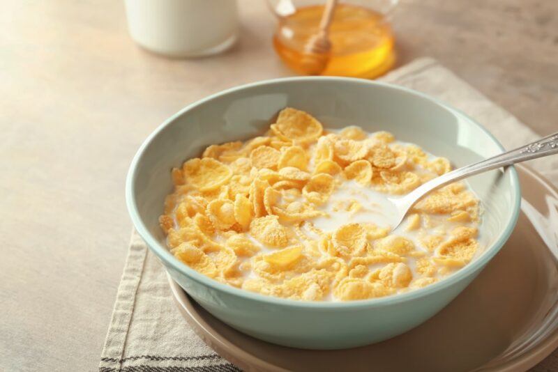 A light colored bowl filled with cornflakes and milk, plus a spoon on a table