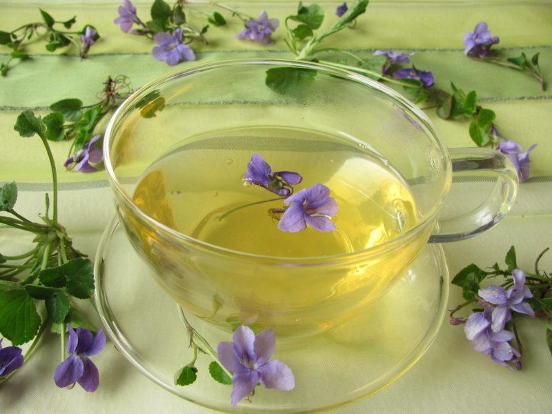 A glass mug that contains light colored tea and a violet. Various violets are scattered around