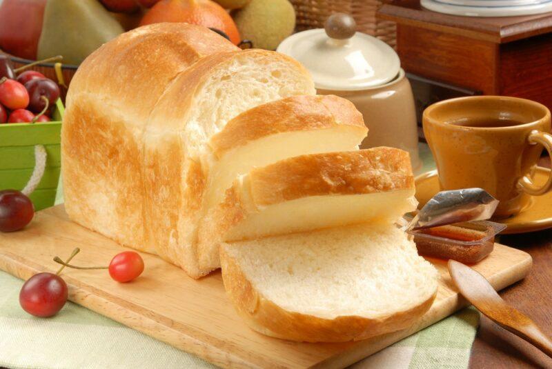 A light colored wooden board with a fresh loaf of bread that's been sliced into thick slices. There's also some fruit, a knife, a small container of honey, a mug of tea, and some kitchen containers.