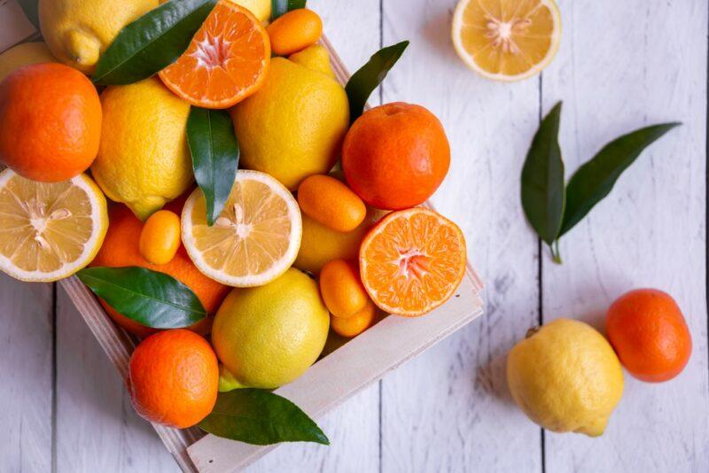 A white table with a white wooden box containing various citrus fruit, including lemons and oranges. Most of these citrus fruit are uncut, but there are a few lemon and orange halves too, along with some dark green leaves