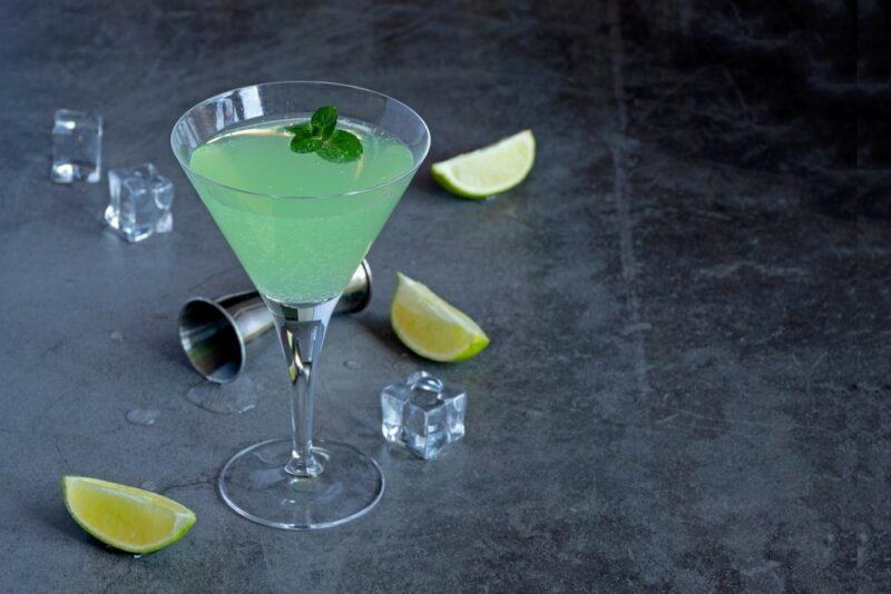 A cocktail glass with a light green tipsy mermaid cocktail on a gray table with a cocktail jigger, some ice, and lime wedges