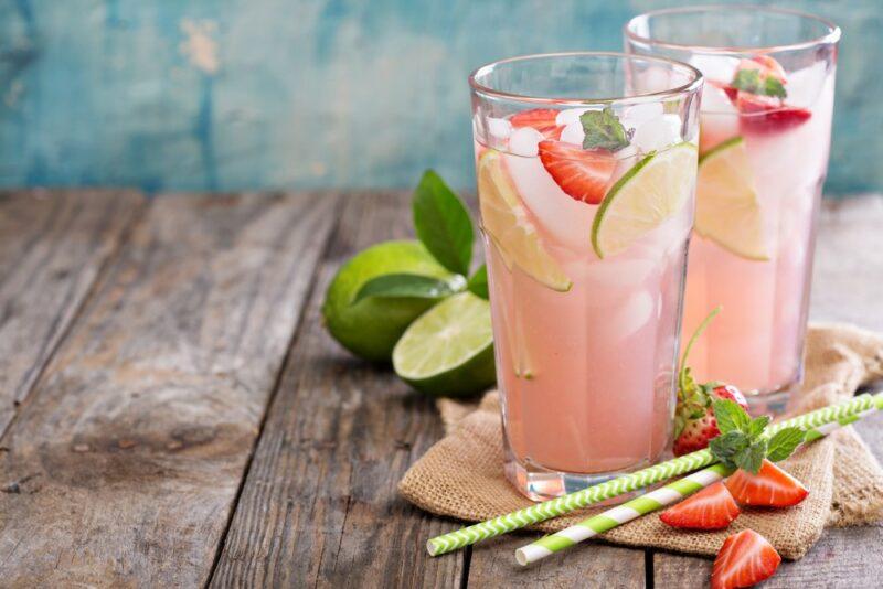 A wooden table with two glasses containing a light pink strawberry and lime energy drink, next to some limes, strawberries, and two straws