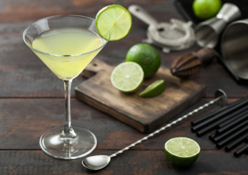 A martini glass containing a lime kamikaze cocktail, next to a cocktail spoon, limes, and bar tools