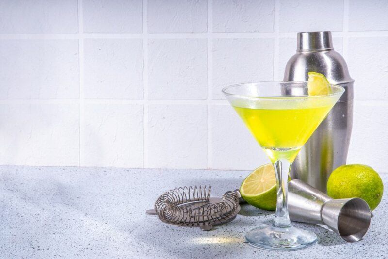 A kitchen bench with a martini glass containing a maladew cocktail, in front of bar equipment, including a cocktail shaker, and some limes