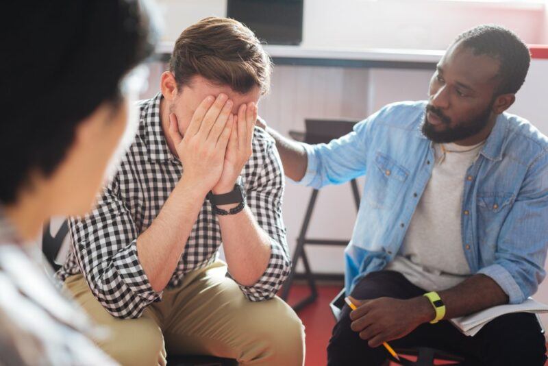 A man crying at some type of support meeting, with a black man comforting him and someone else off to the side