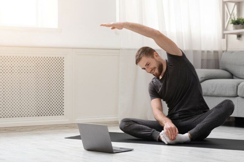 A man practicing yoga at home, sitting in front of his laptop with one arm over his head