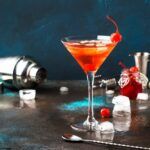 A martini glass containing a Manhattan cocktail on a bar, surrounded by bar tools