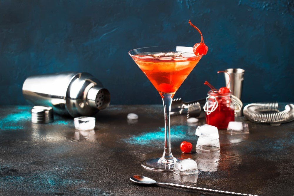 A martini glass containing a Manhattan cocktail on a bar, surrounded by bar tools