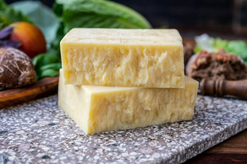 A marble cutting board with two large pieces of aged cheese. There are various vegetables and other ingredients in the background, partly out of focus.