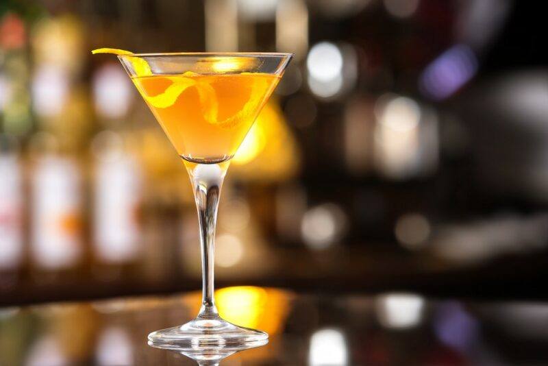 A single glass containing an Agent Orange cocktail on a bar, with out of focus bottles in the background