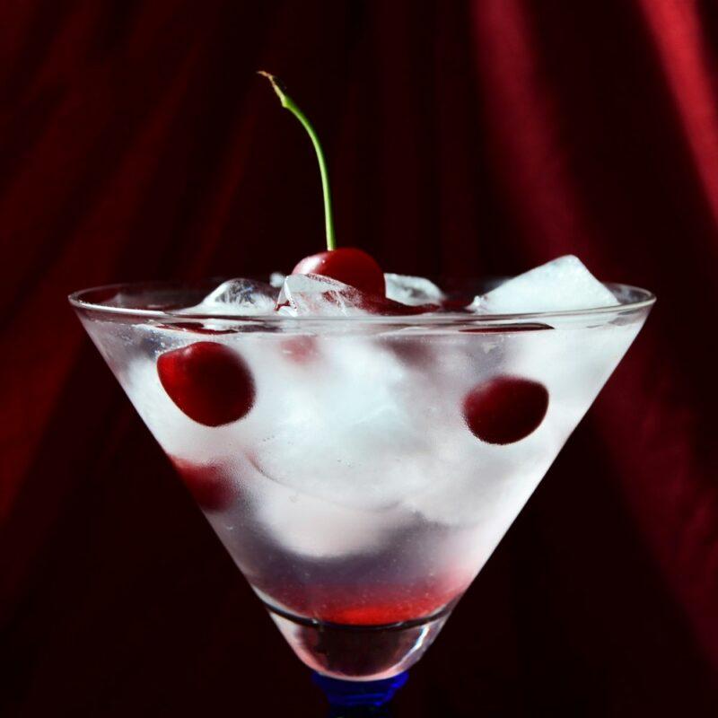 A martini glass containing a black superman cocktail with ice and a cherry